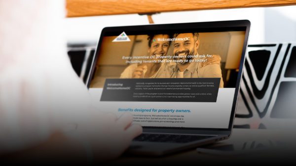 A person is using a laptop to view a website related to property owners. The screen displays information about benefits designed for property owners and features an image of a couple smiling. The laptop is placed on a desk with a wooden texture.