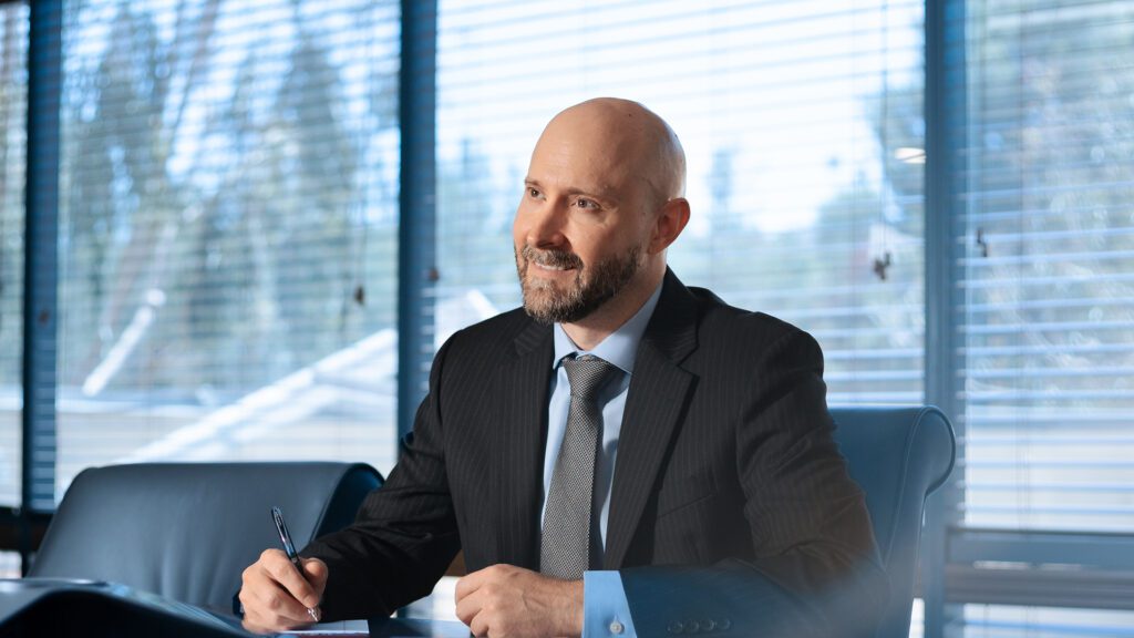 A bald man with a beard, wearing a suit and tie, sits at a table in a bright office with large windows. He is holding a pen and looking off to the side, appearing thoughtful.