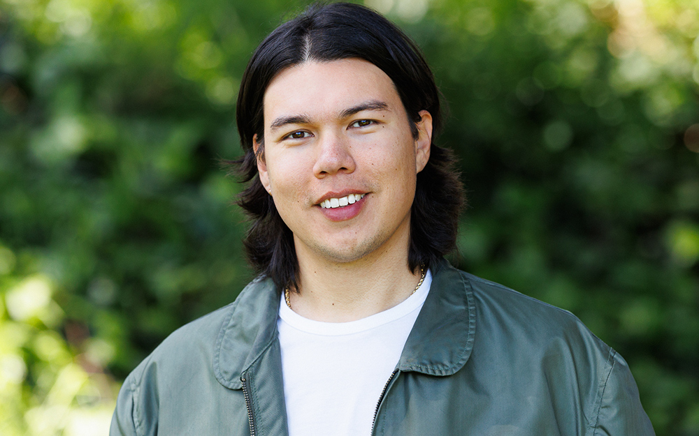 A person with long dark hair, embodying the vibrant energy of a full-service marketing agency, wears a green jacket over a white shirt and smiles outdoors, surrounded by lush greenery.