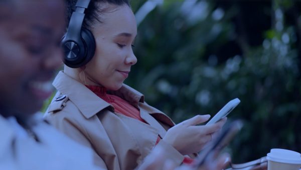 A woman sits outdoors, immersed in emotional content on her smartphone, headphones on and wrapped in a beige trench coat. Beside her, another person partially visible holds a phone and a disposable cup. The scene is framed by lush greenery, suggesting a break amidst the digital marketing hustle.