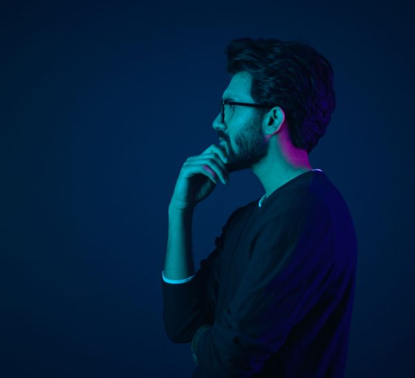 A man with glasses and a beard stands in profile, deep in thought with his hand on his chin, illuminated by blue and purple lighting—perhaps contemplating a new marketing strategy against a dark background.